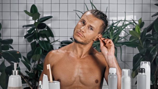 shirtless young man sticking cotton swab in ear near plants on blurred background in bathroom  - Фото, изображение