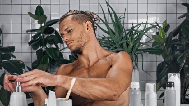 homme torse nu utilisant distributeur de savon près de plantes vertes sur fond flou dans la salle de bain  - Photo, image