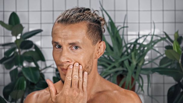 Sakallı ve gömleksiz bir adam banyoda kameraya bakarken yüzüne dokunuyor.  - Fotoğraf, Görsel