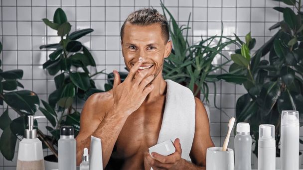 glücklicher Mann, der die Nase im Badezimmer mit Pflanzen auf verschwommenem Hintergrund eincremt - Foto, Bild