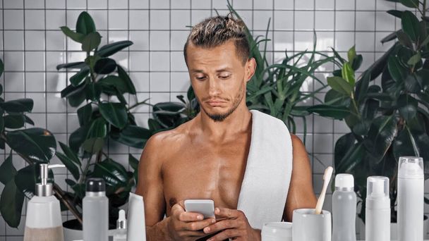 homem sem camisa usando smartphone no banheiro com plantas no fundo borrado - Foto, Imagem