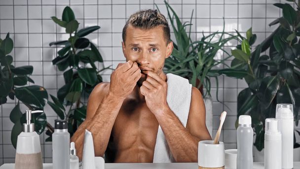 uomo barbuto con asciugamano su denti senza maglietta corpo filo interdentale in bagno vicino a piante su sfondo sfocato - Foto, immagini