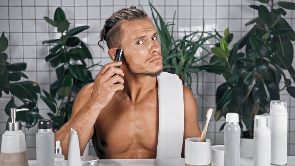 człowiek bez koszuli za pomocą golarki elektrycznej w łazience w pobliżu butelek i roślin na rozmytym tle - Zdjęcie, obraz
