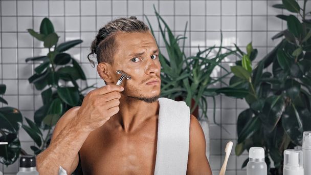 homme torse nu utilisant rasoir dans la salle de bain près des bouteilles et des plantes sur fond flou - Photo, image
