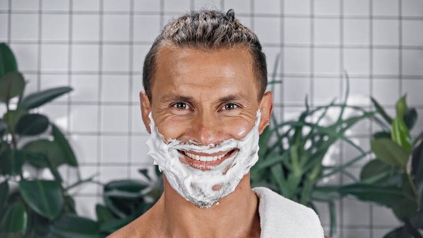 happy man with shaving foam on face looking at camera near plants on blurred background - Photo, Image