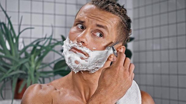 hombre con espuma de afeitar en la cara que sostiene la afeitadora con plantas sobre fondo borroso - Foto, imagen