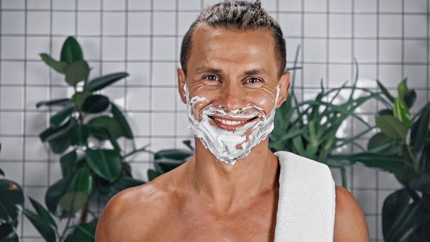 радостный человек с пеной для бритья на лице, смотрящий в камеру рядом с растениями на размытом фоне - Фото, изображение