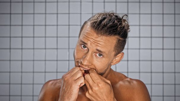 bearded man looking at camera while flossing teeth in bathroom  - Fotoğraf, Görsel