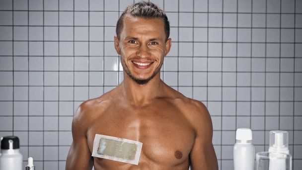 lachende en shirtloze man met wax strip op de borst in de badkamer  - Foto, afbeelding