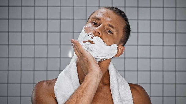 mężczyzna bez koszulki nakładający piankę do golenia na twarz i patrzący w kamerę w nowoczesnej łazience  - Zdjęcie, obraz