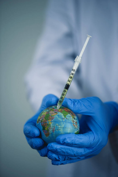 gros plan d'un médecin homme, vêtu d'un manteau blanc et de gants chirurgicaux bleus, vaccinant un globe terrestre - Photo, image
