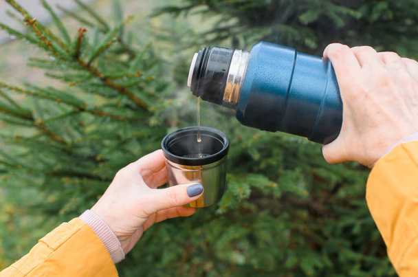Female hands pour hot tea from a thermos close-up, outside in the forest. Senior woman walking and drinking a warm drink in cold weather. - Fotó, kép