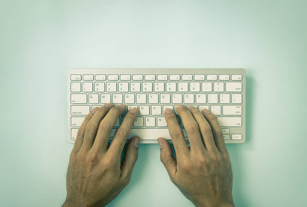 Mano del hombre de negocios escribiendo teclas de teclado de computadora portátil o botón de teclado en la mesa superior o vista superior sobre fondo minimalista pastel azul en tono vintage - Foto, imagen