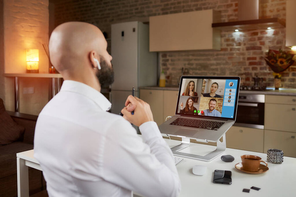 Rückenansicht eines männlichen Angestellten mit verschränkten Armen, der zu Hause auf einem Laptop ferngesteuert an einer geschäftlichen Videokonferenz arbeitet. Ein Glatzkopf mit Bart und Kopfhörer bei einem Online-Treffen mit seinen Kollegen - Foto, Bild