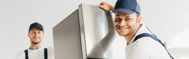 cheerful indian mover in uniform moving fridge near coworker on blurred background, banner - Photo, image