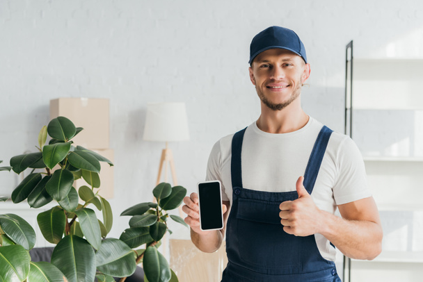 cheerful mover in overalls holding smartphone with blanks screen and showing thumb up  - Fotografie, Obrázek