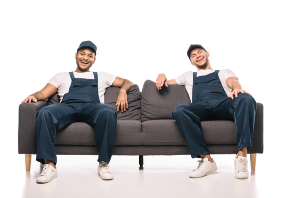 full length of cheerful multicultural movers laughing while sitting on sofa on white - Photo, Image
