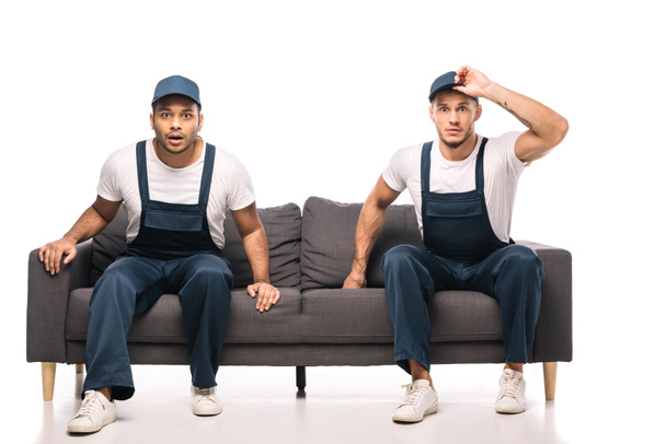 full length of shocked mover adjusting cap while sitting on sofa with indian worker on white - Foto, Imagen