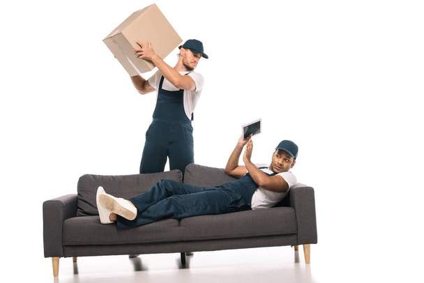 напуганный индийский работник держит цифровой планшет, опираясь на диван и жестикулируя рядом с сердитым мовером с коробкой на белом - Фото, изображение