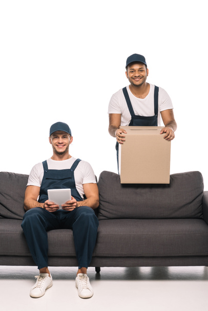 χαρούμενα εργαζόμενος κάθεται στον καναπέ με ψηφιακή ταμπλέτα κοντά indian mover κρατώντας κουτί σε λευκό - Φωτογραφία, εικόνα