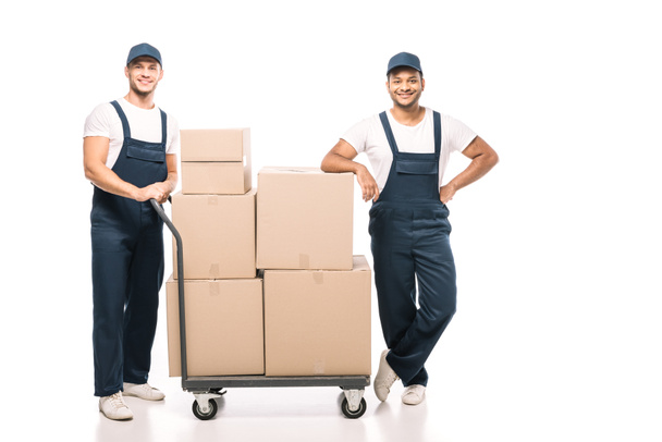 full length of joyful multicultural movers in uniform and caps standing near hand truck with carton boxes on white - Фото, изображение