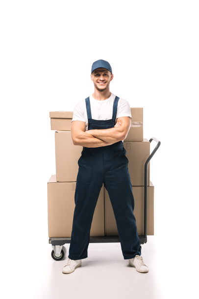 full length of cheerful mover in uniform and cap standing with crossed arms near hand truck with carton boxes on white - Foto, Bild