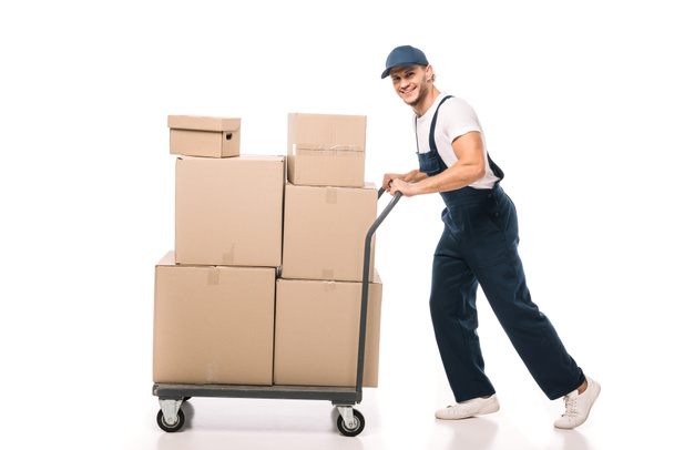 full length of cheerful mover in uniform and cap pulling hand truck with carton boxes on white - Photo, Image