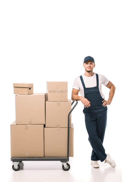 full length of cheerful mover in uniform and cap leaning on hand truck with carton boxes on white - Foto, immagini