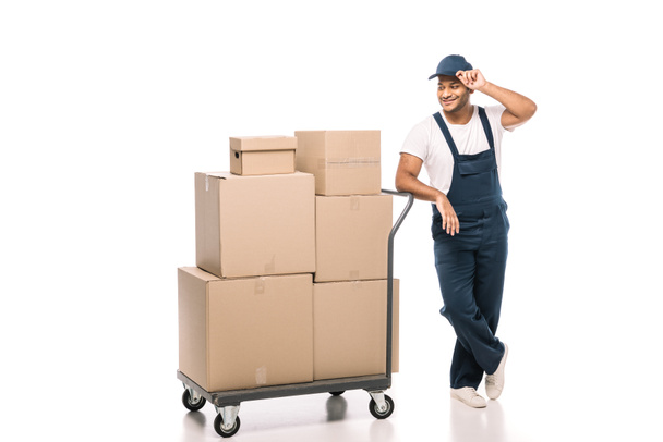 joyful indian mover standing near hand truck with carton boxes and adjusting cap on white - Photo, Image