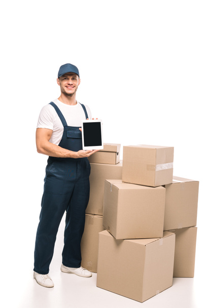 full length of cheerful mover in uniform and cap holding digital tablet with blank screen near carton boxes on white - Фото, изображение