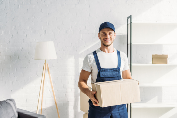 cheerful mover in cap holding carton box in apartment  - Photo, Image