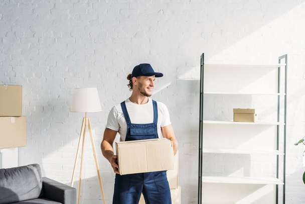 happy mover in cap holding carton box and looking away in apartment  - Photo, Image