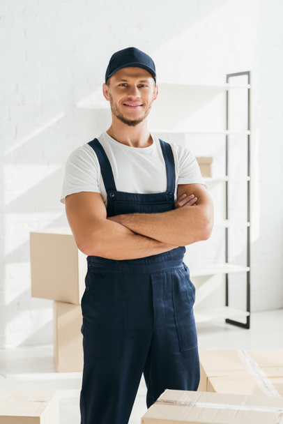 happy mover in uniform and cap standing with crossed arms in apartment  - Photo, Image