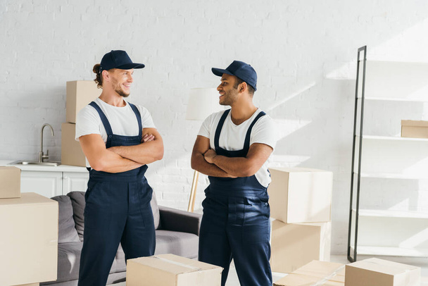 cheerful movers in caps standing with crossed arms while looking at each other near boxes in room - Photo, Image