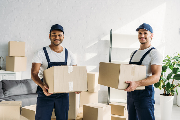 cheerful multiethnic movers in caps and uniform carrying boxes in apartment  - Photo, Image