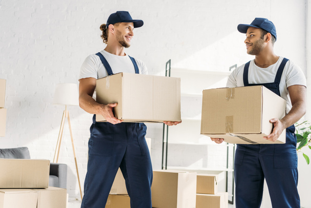 smiling multiethnic movers in caps and uniform carrying boxes in apartment  - Photo, Image