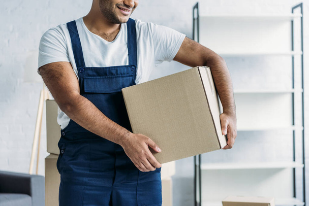 cropped view of cheerful indian courier in uniform holding box in apartment  - Foto, Bild