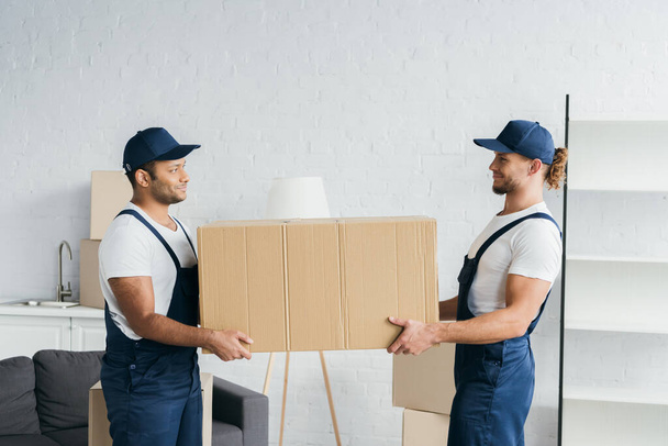 side view of multiethnic movers looking at each other and holding carton box  - Foto, Bild