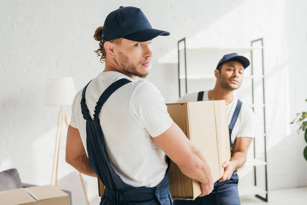 multiethnic movers in uniform and caps carrying huge carton box  - Photo, Image