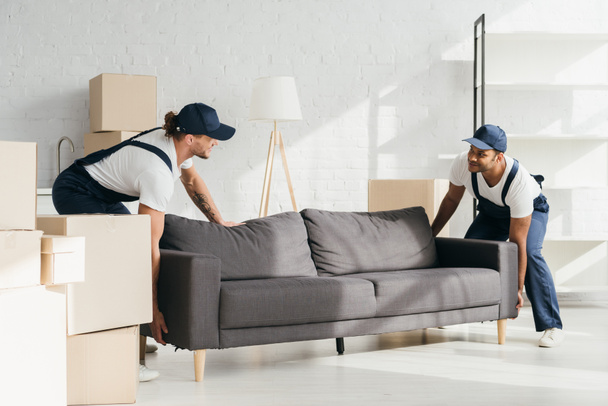 happy multicultural movers in uniform smiling while carrying couch in apartment - Фото, изображение