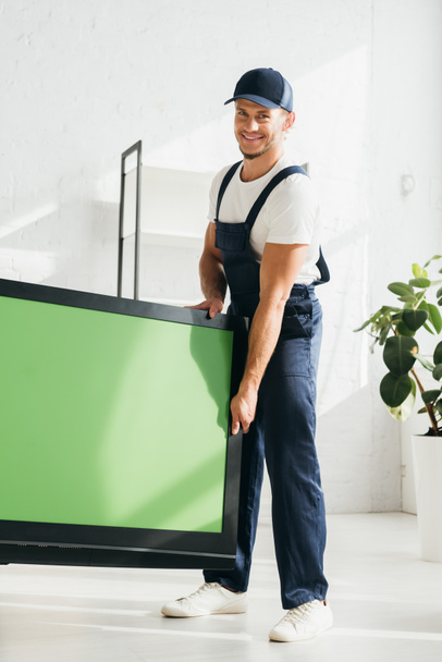 sonriente mover en uniforme llevando plasma tv con pantalla verde en apartamento  - Foto, Imagen