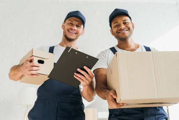 low angle view of cheerful multicultural movers smiling while looking at camera and holding boxes in apartment  - Photo, Image