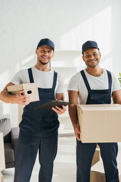 cheerful multicultural movers smiling while looking at camera and holding boxes in apartment  - Photo, Image