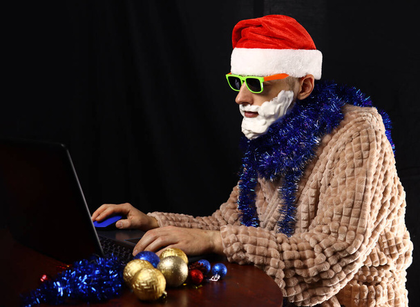 Santa mal loco en gafas de sol, barba de espuma en la ropa de casa sentado en un ordenador portátil - Foto, imagen