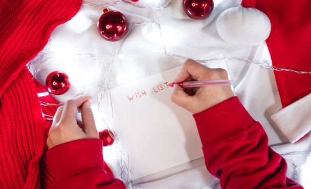 Escribe una lista de deseos para Santa con un bolígrafo rojo en una hoja blanca en blanco. Bolas rojas de Navidad, sombrero de Santa y bufanda hecha a mano. Escribe una carta con deseos en una manta blanca. - Foto, imagen