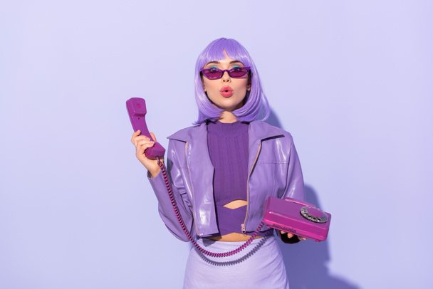 здивована молода жінка, одягнена в ляльковий стиль з ретро телефоном на фіолетовому барвистому фоні
 - Фото, зображення