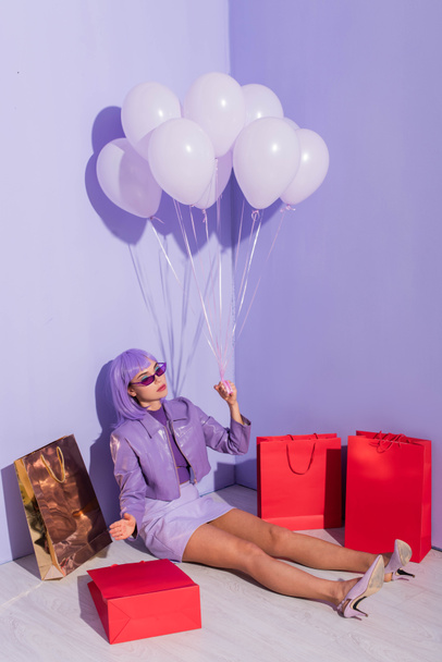 mujer joven vestida con estilo de muñeca sentada con bolsas de compras y globos sobre fondo de color violeta - Foto, imagen