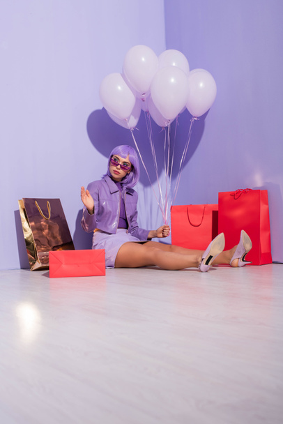 молодая женщина одета в кукольный стиль сидя с пакетами покупок и воздушными шарами на фиолетовом красочном фоне - Фото, изображение
