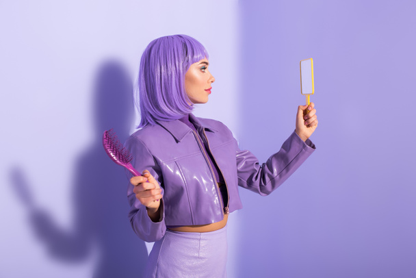 young woman dressed in doll style with hair brush and mirror on violet colorful background - Foto, afbeelding