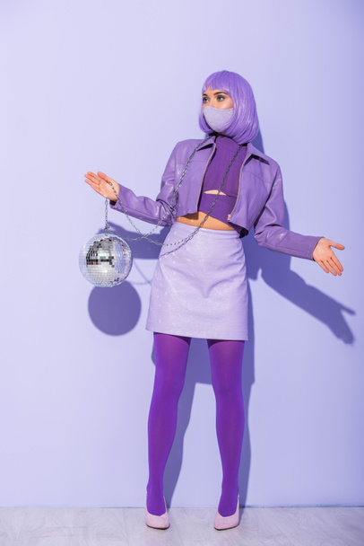 young woman dressed in doll style in medical mask with disco ball on violet colorful background - Fotó, kép